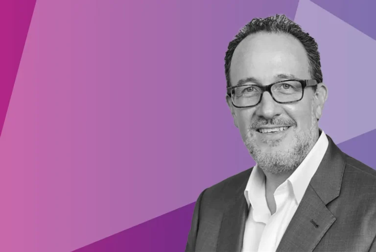 Smiling man with glasses and a beard wearing a suit jacket and white shirt, in front of a geometric purple background.
