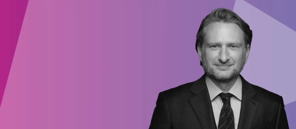 A man in a suit and tie is on the right side of the image, with a purple and pink geometric background on the left.