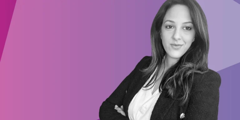 A woman with long dark hair, wearing a blazer and blouse, stands with arms folded, smiling slightly. The background features purple and pink gradient geometric shapes.