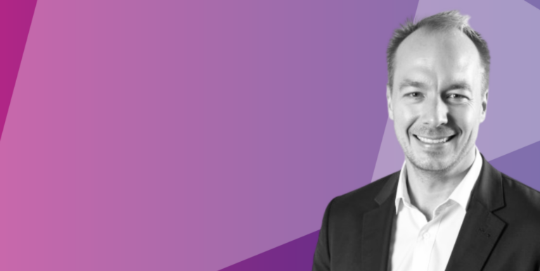 A man in a suit smiles, positioned at the lower right of a purple geometric background. The image is mostly purple with the man shown in black and white.