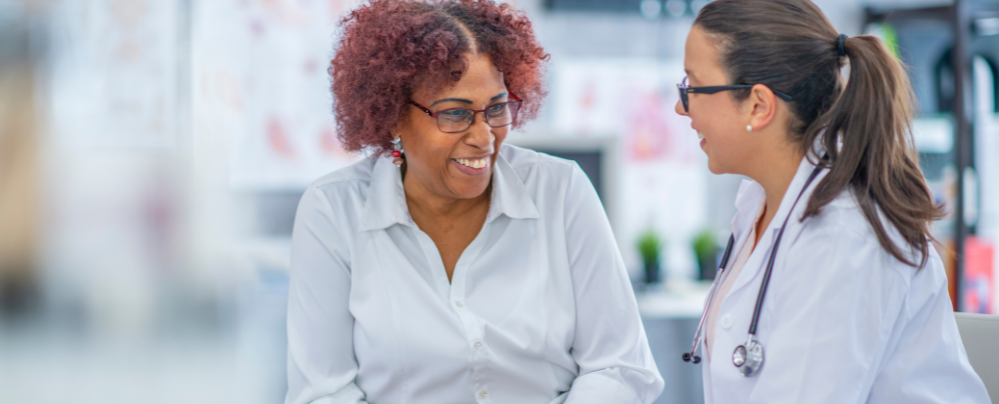 A doctor with a stethoscope talks and smiles with a patient in a consultation room, both appearing engaged and happy during the conversation.