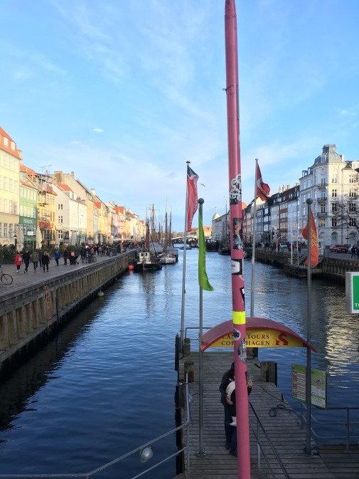 A canal with boats moored along the sides, colourful buildings lining both banks, and people walking by. A pink pole and a small tour kiosk are in the foreground under a clear blue sky.