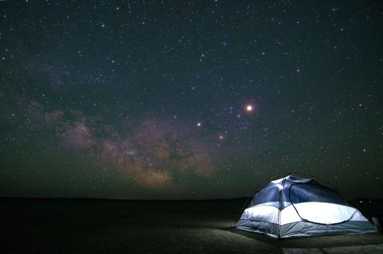 A glowing tent sits on open ground at night under a vast, star-filled sky with the Milky Way visible in the background.