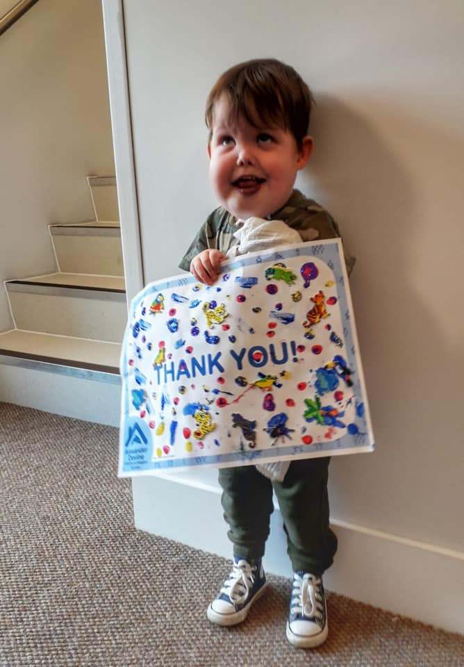 A young child smiles whilst holding a colourful, decorated sign that says THANK YOU! The child stands indoors near a staircase, wearing green trousers, a shirt, and trainers.