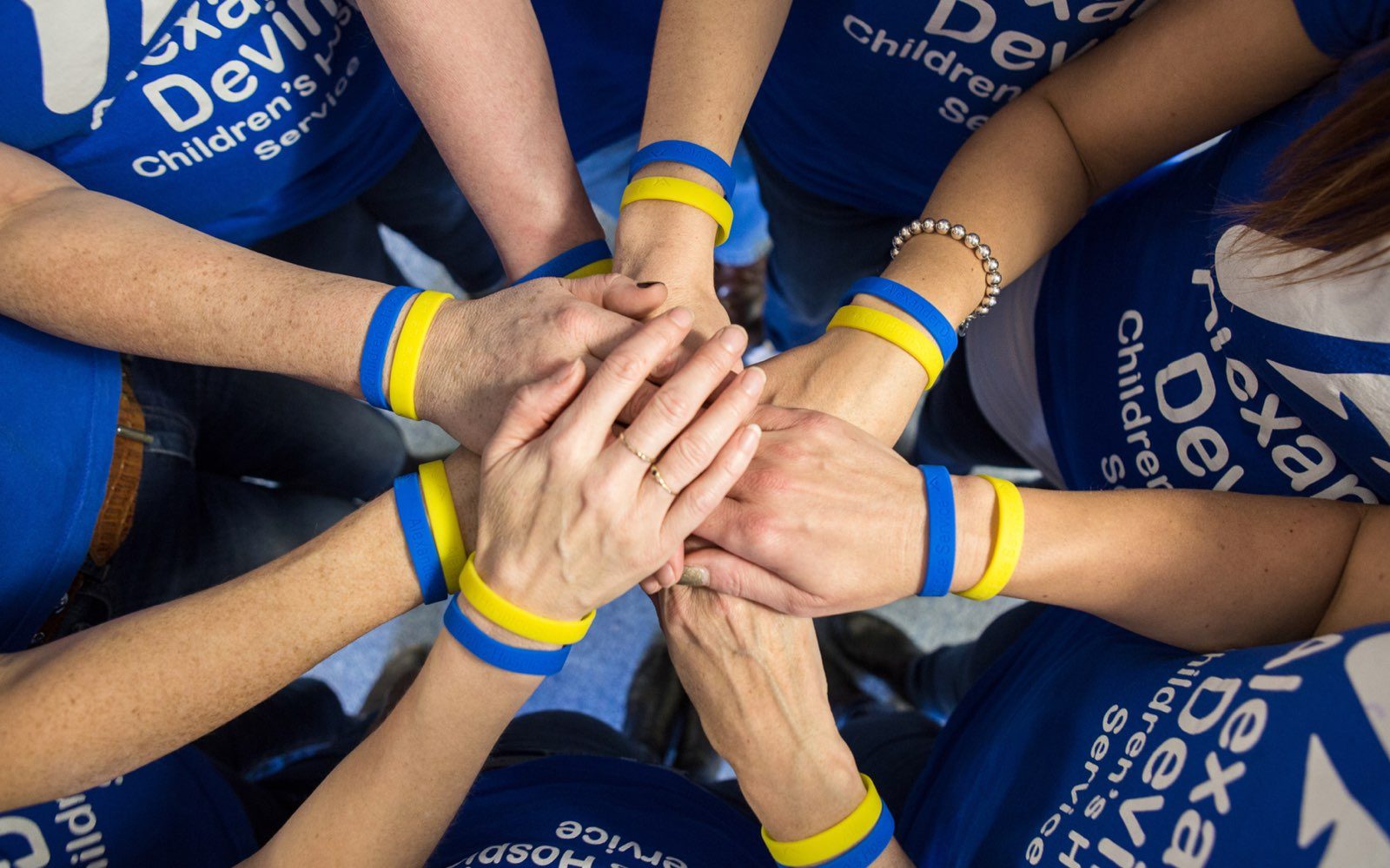 A group of people wearing blue shirts and blue and yellow wristbands join their hands together in a circle, symbolising unity and teamwork.