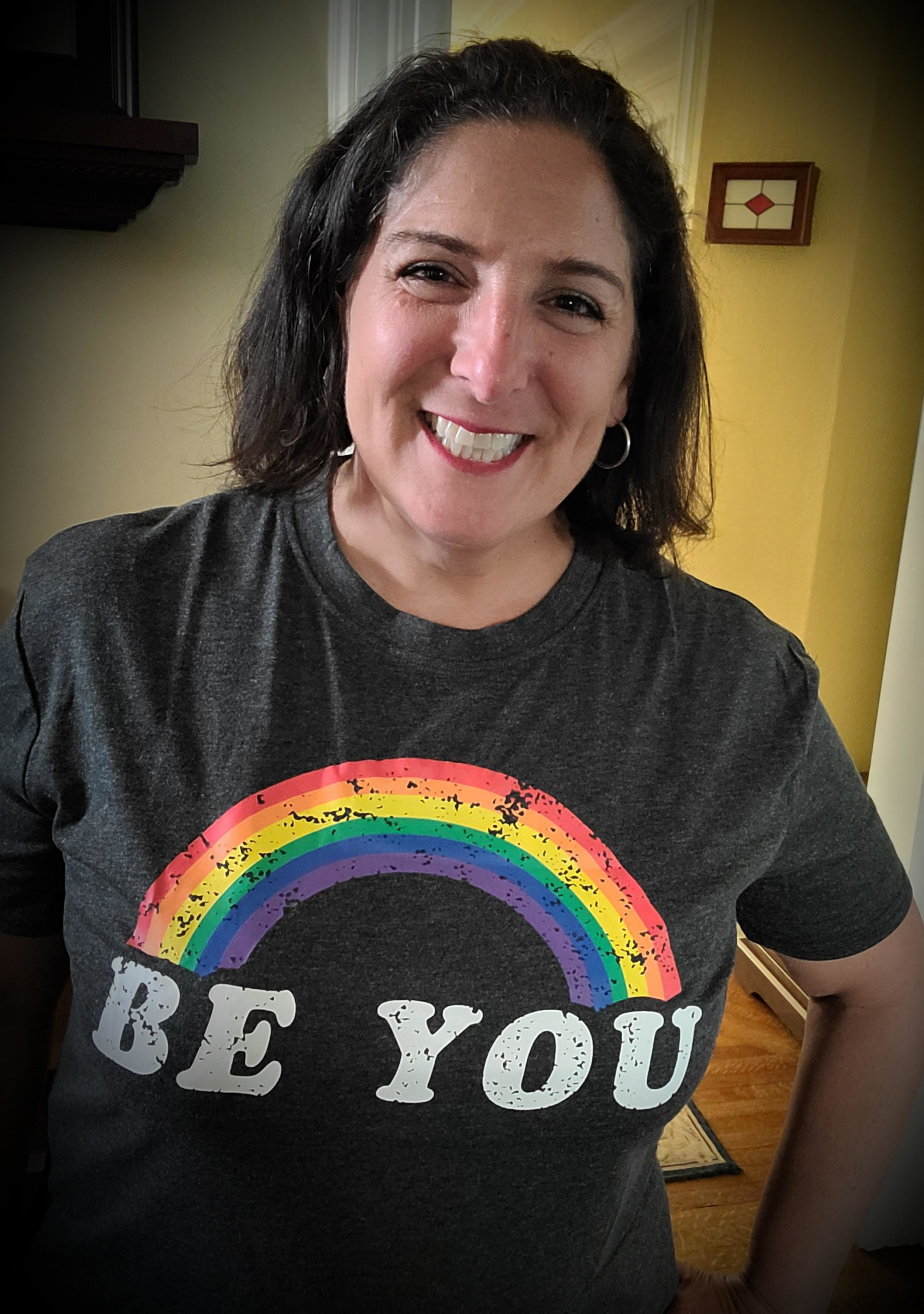 A woman smiles, wearing a dark T-shirt with a rainbow and the words BE YOU in bold white letters. She has medium-length dark hair and stands indoors with a light yellow wall behind her.