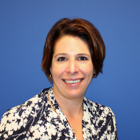 Smiling woman with short brown hair wearing a patterned blouse, standing in front of a plain blue wall.