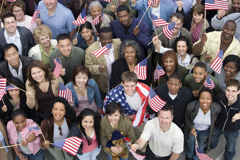 A diverse group of people smile and wave American flags, standing close together and looking up at the camera, celebrating patriotism and unity.