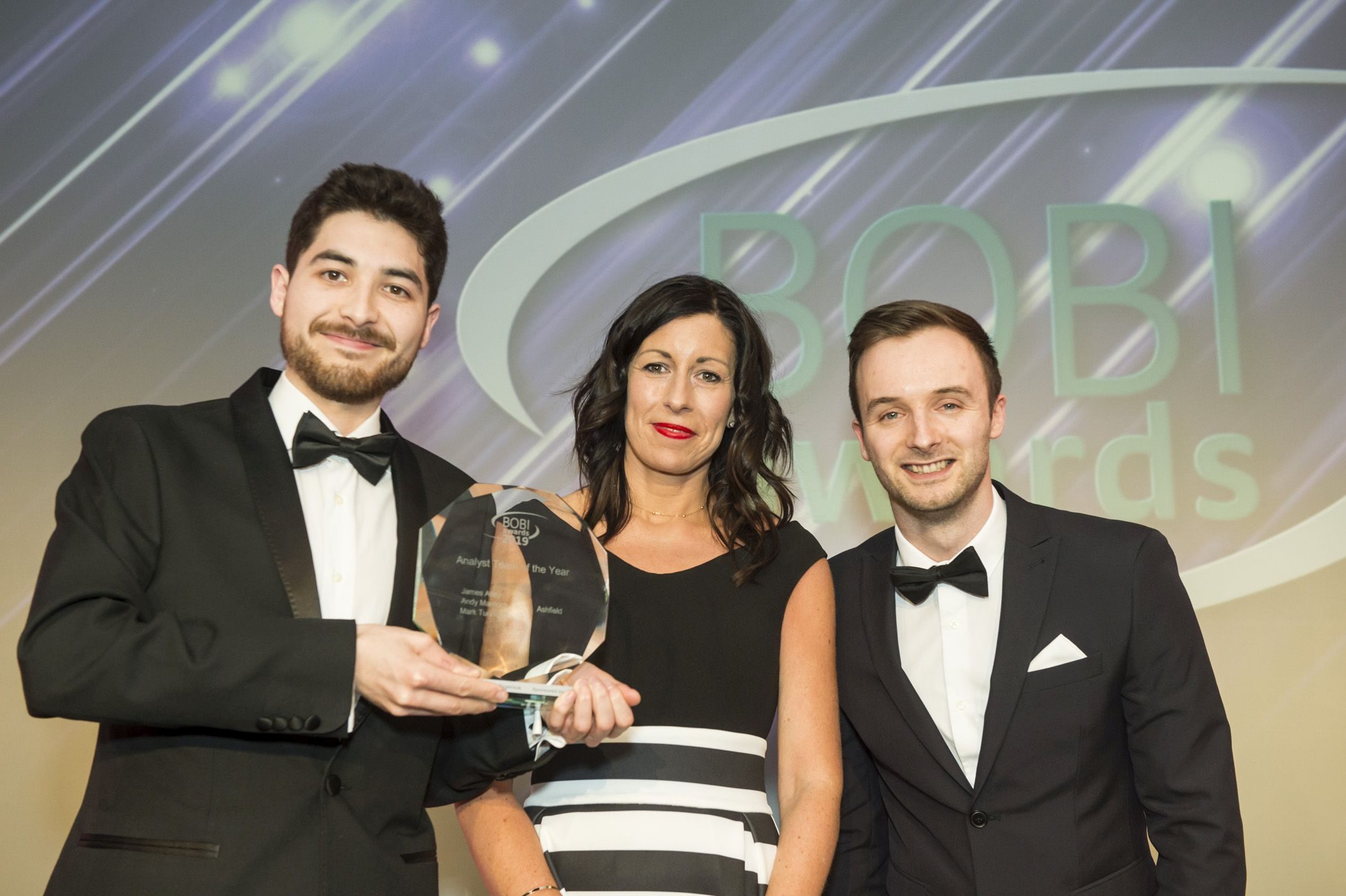 Three people in formal attire pose on stage; the man on the left holds a glass award. The background displays the text BOBI Awards on a lit backdrop.