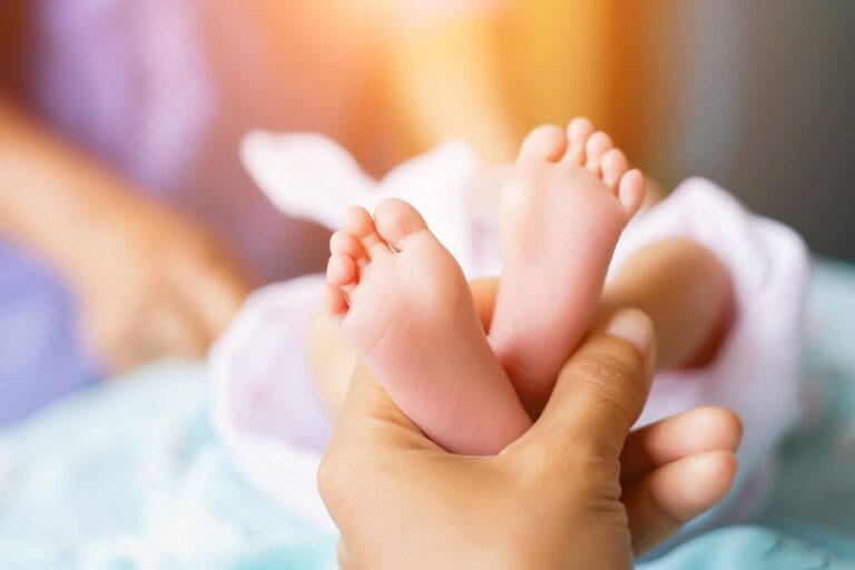A close-up of an adult hand gently holding the tiny feet of a baby, with a soft, warm light in the background.
