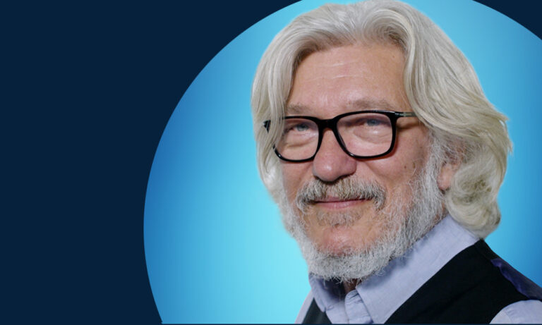 An older man with white hair, a beard, and glasses smiles at the camera. He wears a light blue shirt and dark waistcoat. The background has a dark and light blue gradient.