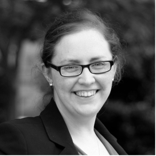 A woman with glasses and dark hair tied back smiles at the camera. She is wearing a dark blazer and earrings. The background is blurred and appears to be outdoors. The photo is in black and white.