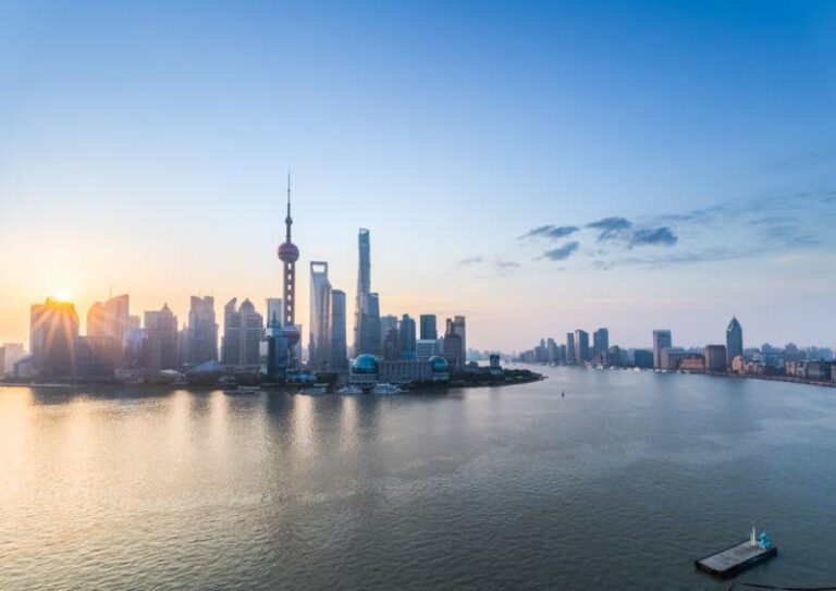 Shanghai skyline at sunrise, with modern skyscrapers and the Oriental Pearl Tower by the Huangpu River, under a clear blue sky.