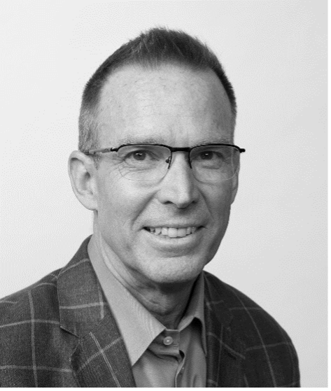 Black and white portrait of a man wearing glasses, a chequered blazer, and a collared shirt, smiling at the camera against a plain background.