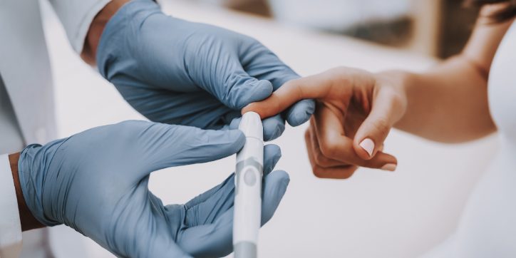 A healthcare professional wearing blue gloves uses a small device to prick a patient's finger, likely for a blood test or glucose check.