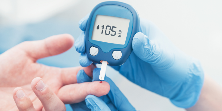 A person in blue gloves uses a blood glucose metre on someone's finger, displaying a reading of 105 mg/dL.