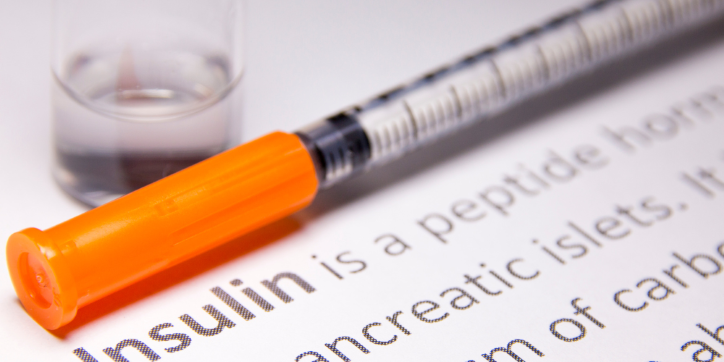 A syringe with an orange cap is lying on a sheet of paper with the word Insulin in bold text, next to a small clear container.