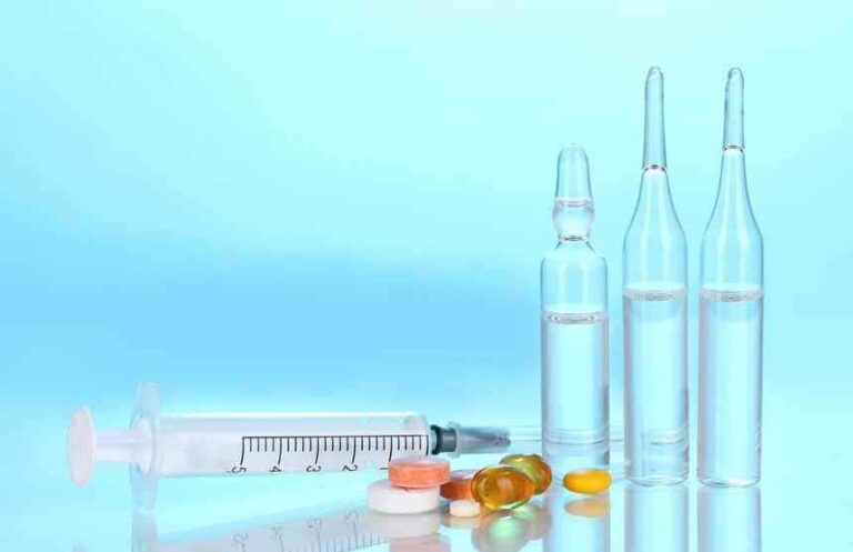 A syringe, three glass ampoules filled with clear liquid, and various pills and capsules are arranged on a reflective surface against a light blue background.