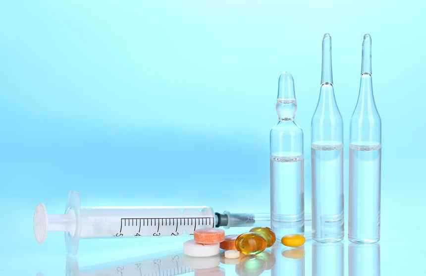 A syringe, three glass ampoules filled with clear liquid, and various pills and capsules are arranged on a reflective surface against a light blue background.