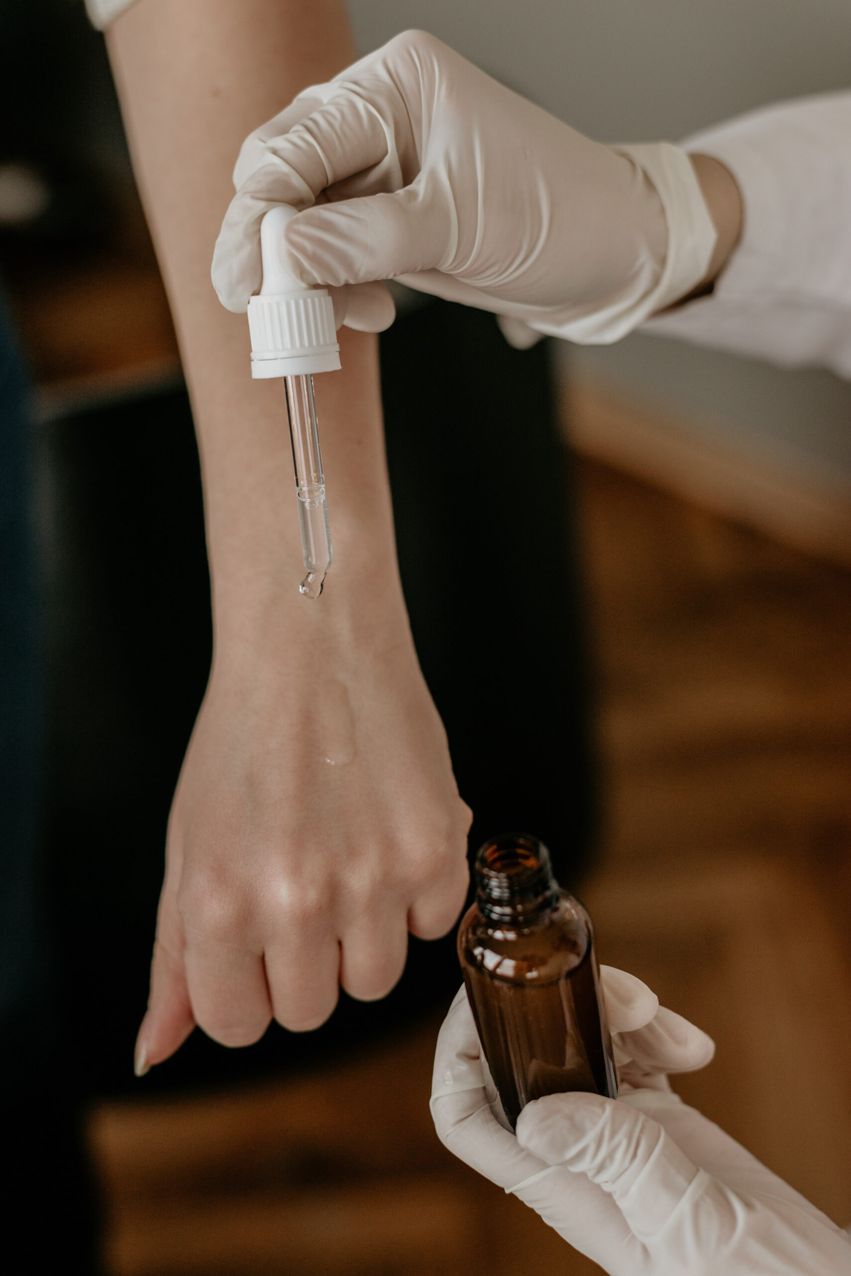 A gloved hand uses a dropper to apply a clear liquid onto the back of another person's hand, with a brown glass bottle held below.