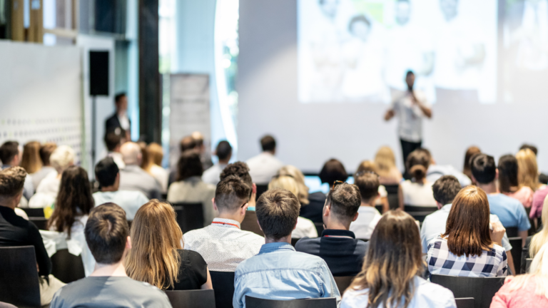 A group of people sit facing a presenter speaking in front of a large screen in a bright, modern conference room.