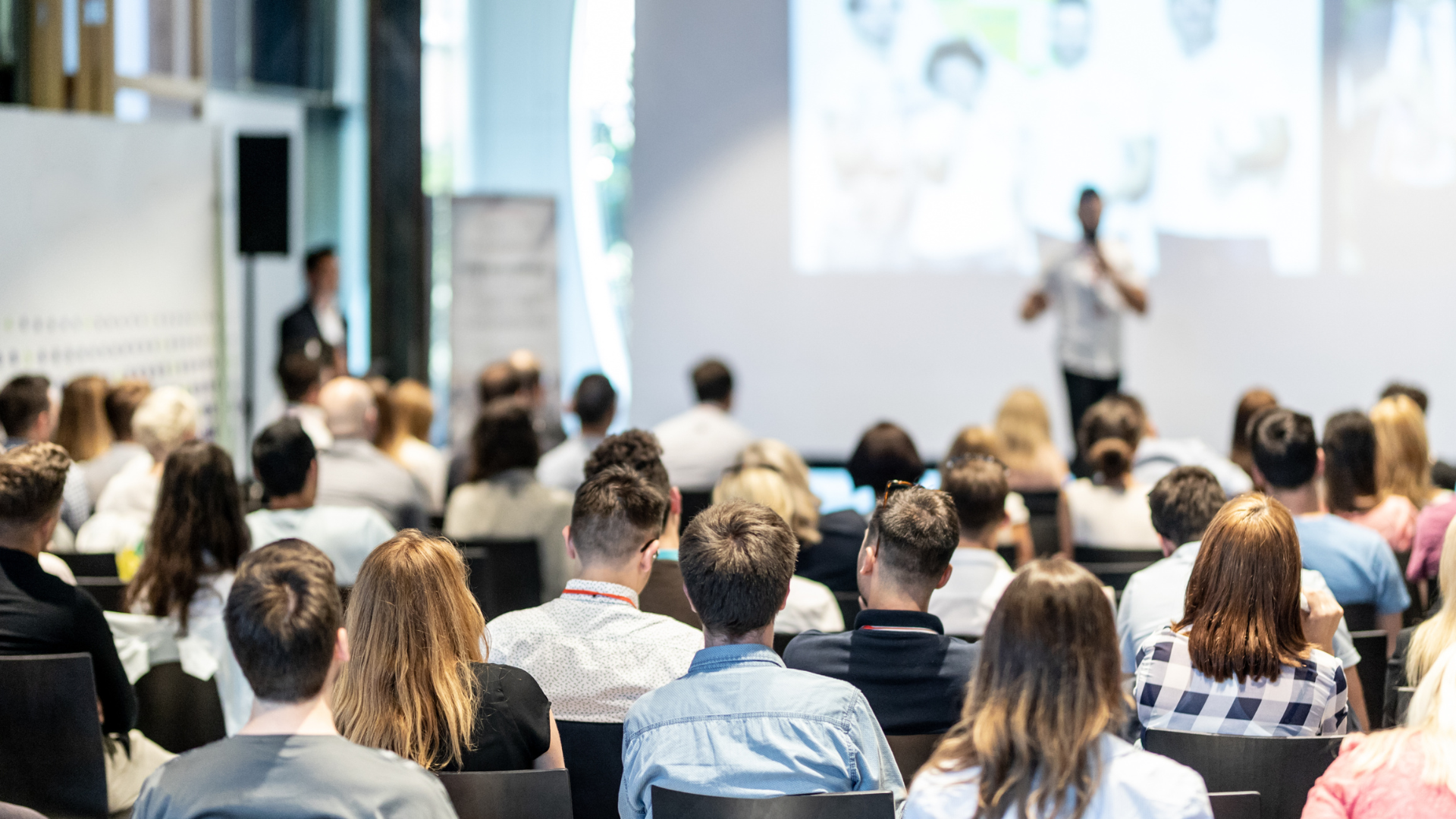A group of people sit facing a presenter speaking in front of a large screen in a bright, modern conference room.