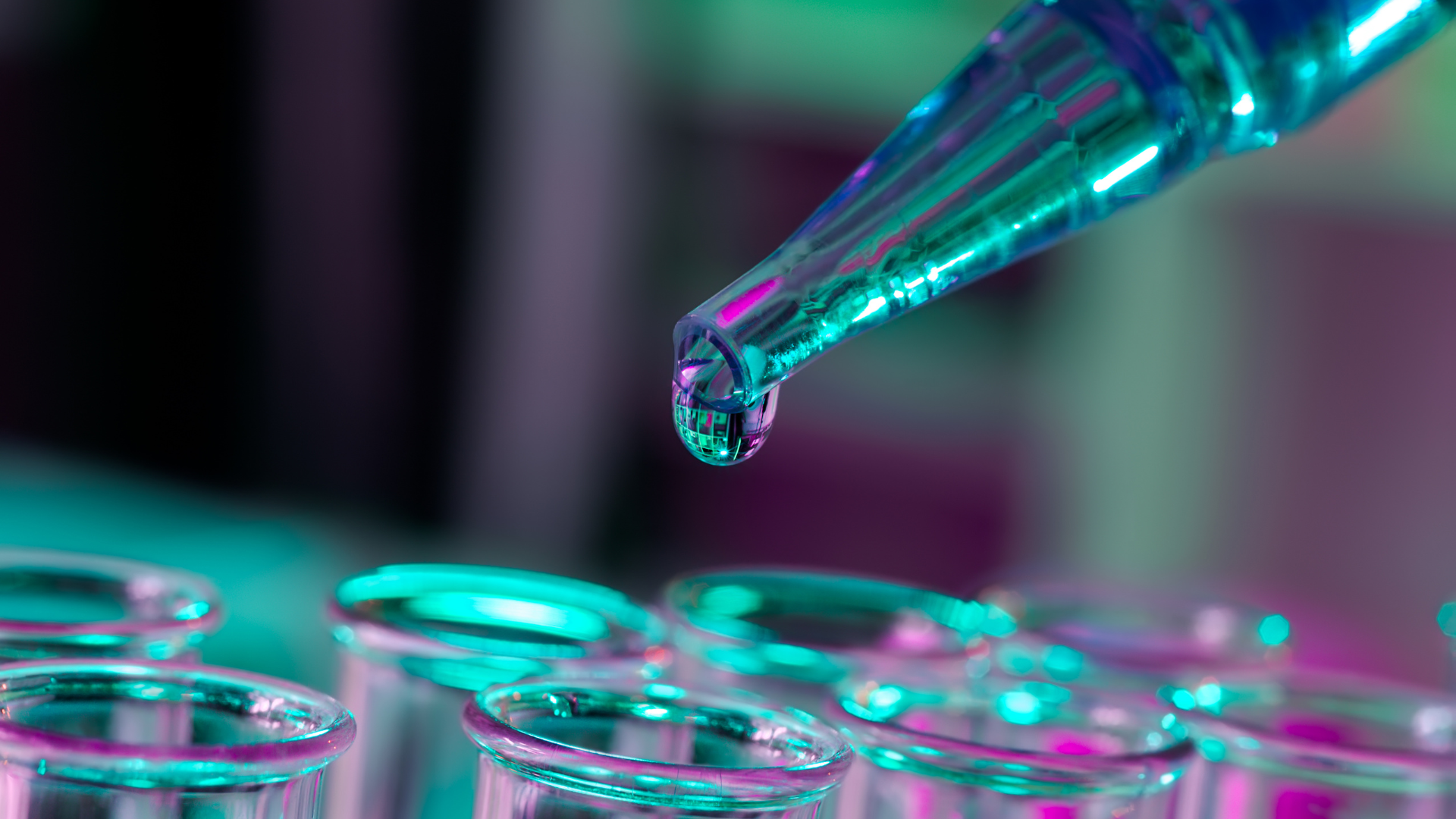 A close-up of a pipette releasing a drop of liquid into one of several glass test tubes, illuminated by blue and purple lighting.