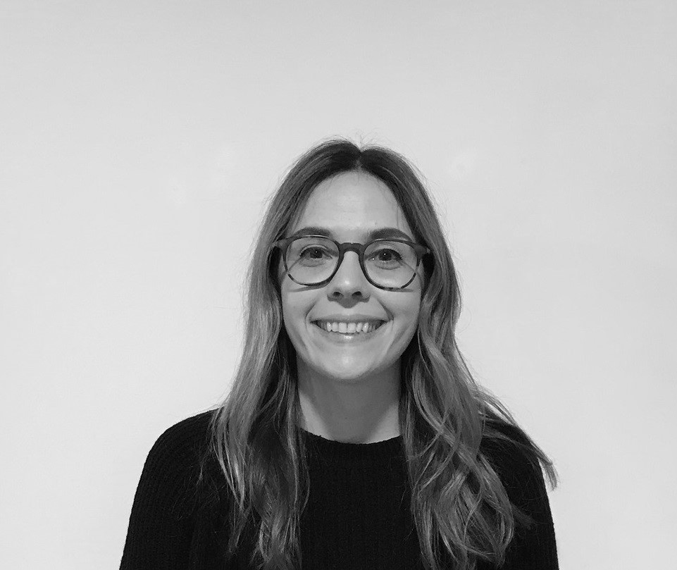 A woman with long hair and glasses smiles at the camera. She is wearing a dark top and is in front of a plain light-coloured background. The image is in black and white.