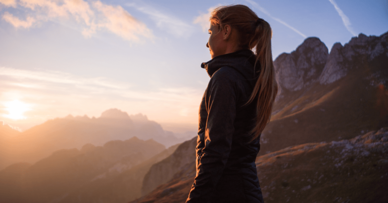 A woman with a ponytail stands on a mountain at sunset, looking out at a scenic view of peaks and a glowing sky.