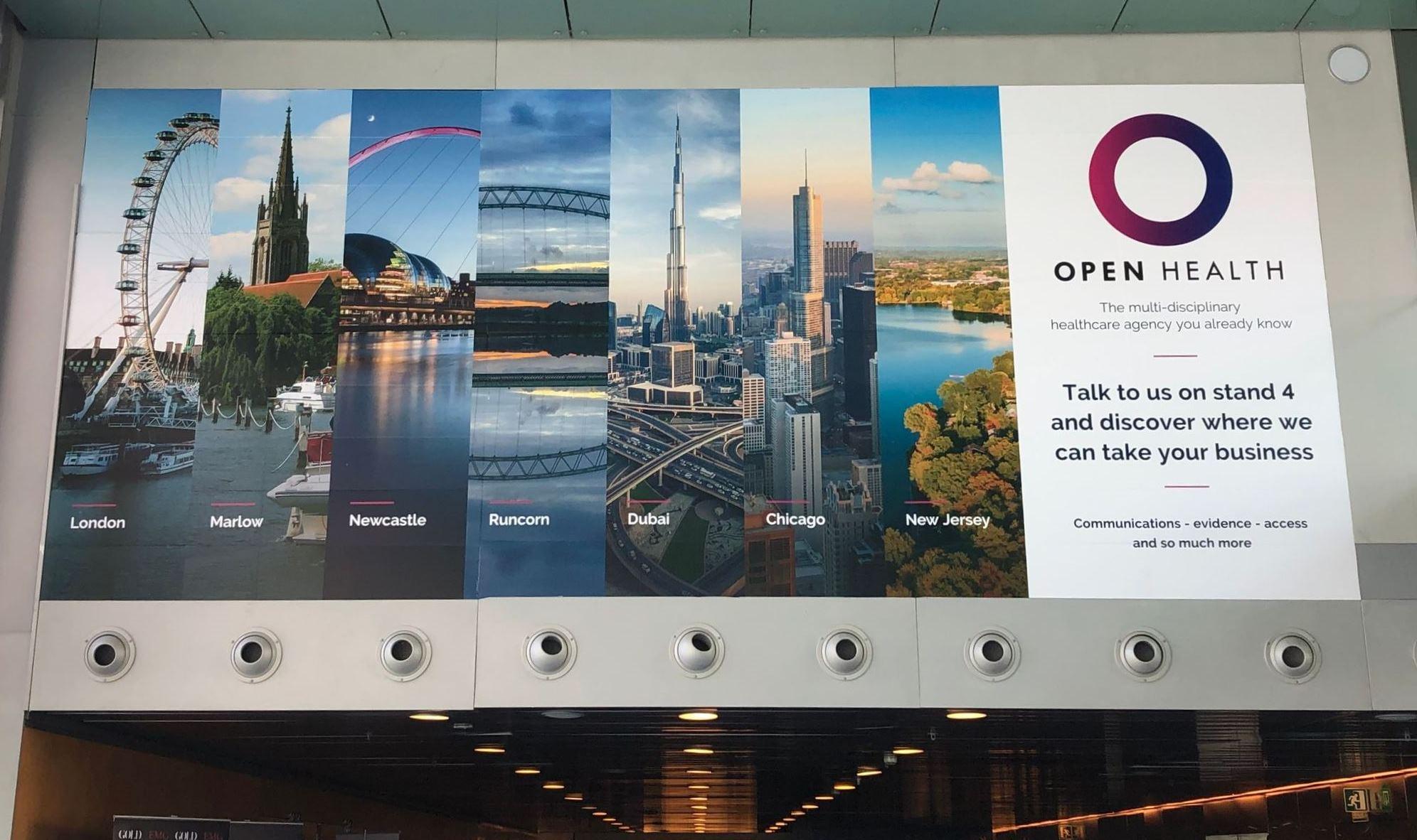 A banner for OPEN Health shows cityscapes of London, Marlow, Newcastle, Runcorn, Dubai, Chicago, and New Jersey, inviting people to visit their stand and learn more about their healthcare agency services.