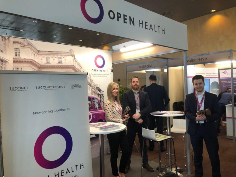 Three people stand and smile at an OPEN Health stand at a conference. The stand displays the company logo and marketing banners. There are tables, chairs, and other stand displays in the background.
