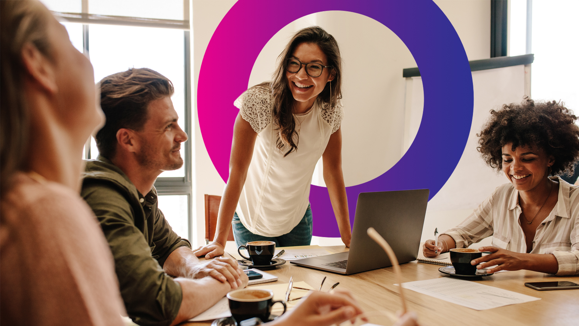 Four people sit around a table with coffee and a laptop, engaged in a lively discussion, whilst one woman stands and smiles at the group. A large circular graphic is on the wall behind her.