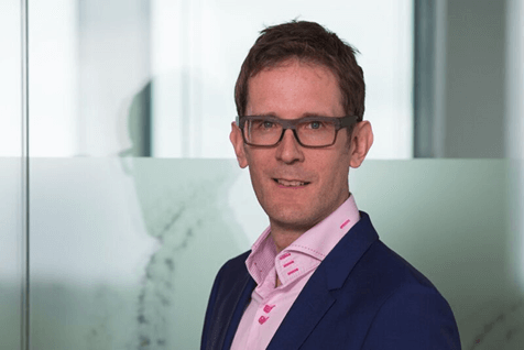 Man with short brown hair and glasses wearing a dark blue blazer and light pink shirt, standing indoors in front of a frosted glass wall.