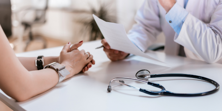 A doctor and a patient sit opposite each other at a desk. The doctor is holding a piece of paper. A stethoscope is on the table between them. The patient's hands are clasped, and both people are partially visible.