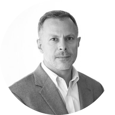 Black and white portrait of a middle-aged man with short hair, wearing a suit jacket over a collared shirt, looking directly at the camera, against a plain white background.