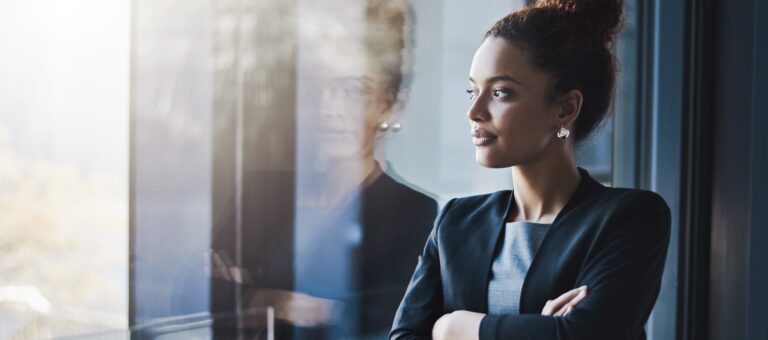 A woman in business attire stands with arms folded, looking thoughtfully out of a window, with her reflection visible on the glass.