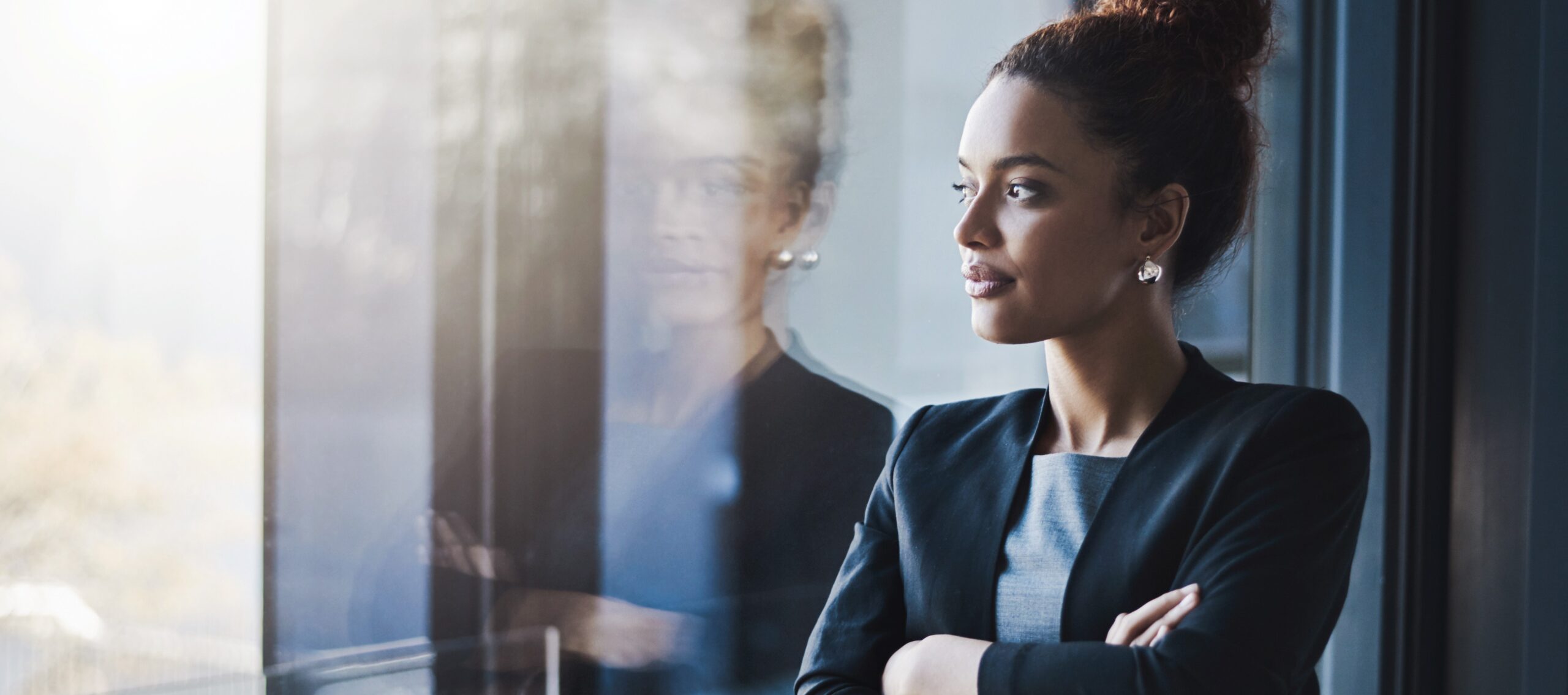 A woman in business attire stands with arms folded, looking thoughtfully out of a window, with her reflection visible on the glass.