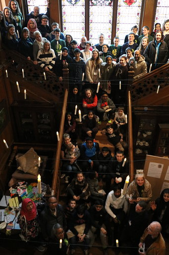 A large, diverse group of people pose on a staircase and landing in a building with stained glass windows, looking up at the camera. There are hanging lights, and some people hold objects or wear fancy dress.