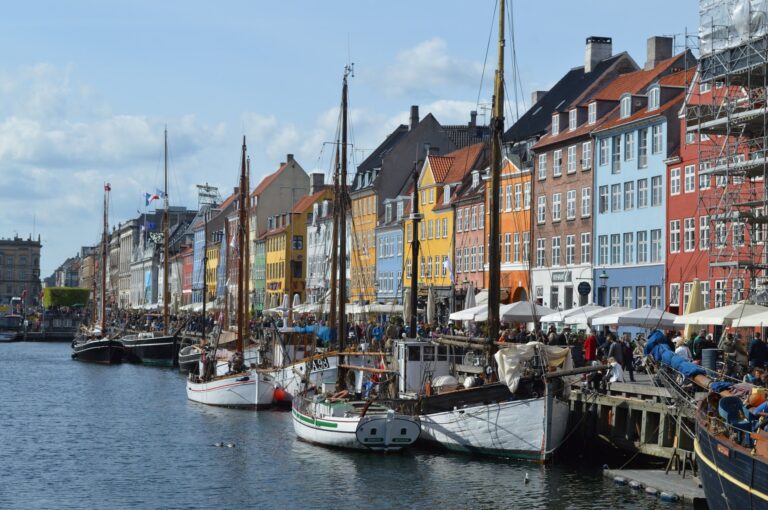 Colourful buildings line a busy canal with docked boats and people walking along the waterfront under a partly cloudy sky.