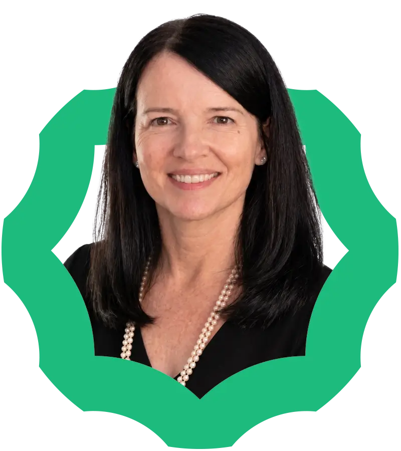 A woman with long dark hair wearing a black top and a pearl necklace, smiling against a green, decorative frame on a white background.