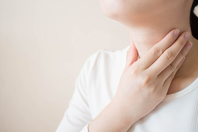 A person touching their throat with one hand, possibly indicating discomfort or a sore throat, against a neutral background.