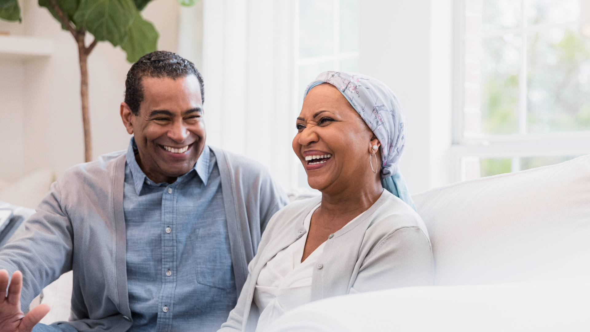 A smiling older man and woman sit together on a white sofa in a bright living room. The woman wears a headscarf, and both appear happy and relaxed as they laugh together.