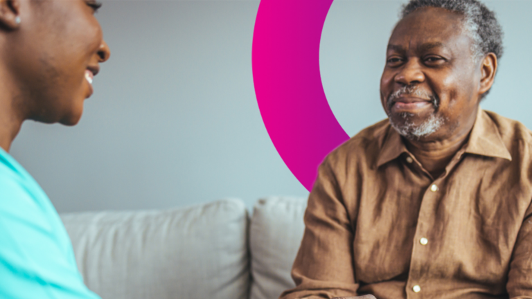 An older man in a brown shirt smiles while sitting on a sofa, talking to another person. A pink semi-circle design appears on the wall in the background.