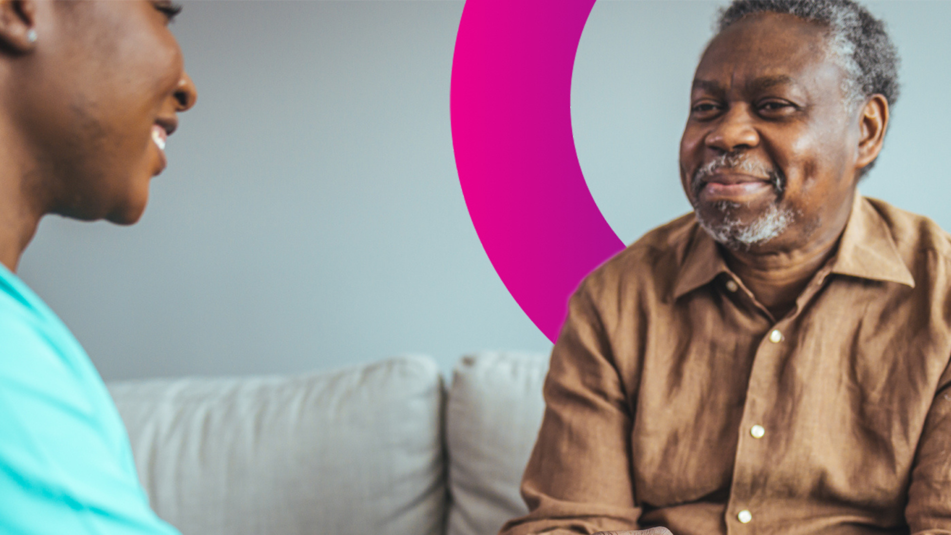 An older man in a brown shirt smiles while sitting on a sofa, talking to another person. A pink semi-circle design appears on the wall in the background.