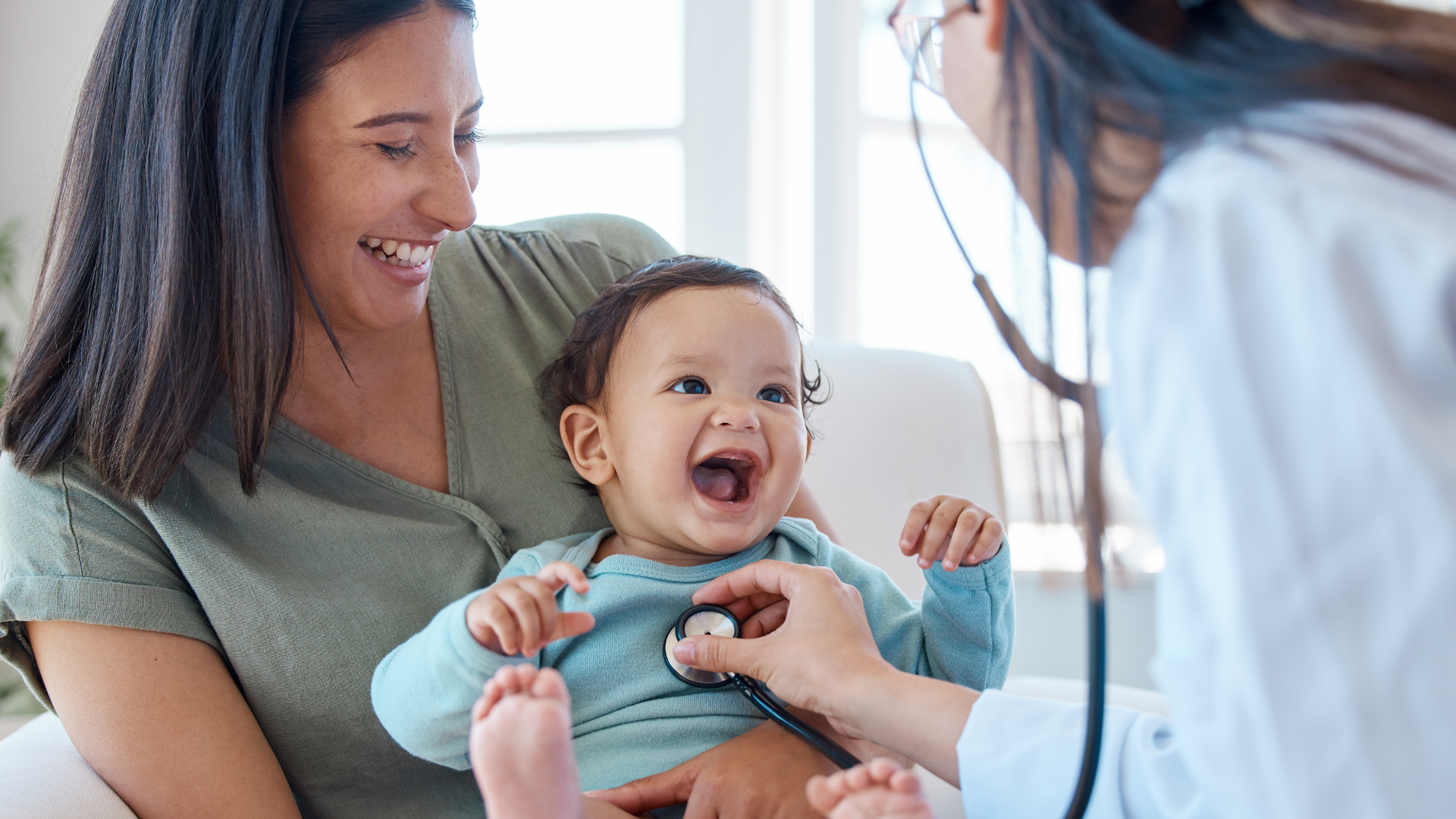 A smiling mother holds her laughing baby while a doctor uses a stethoscope to check the baby's chest during a medical examination in a bright room.
