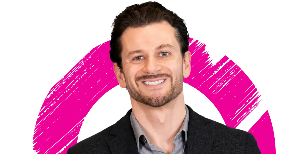 A man with dark hair and a beard, wearing a dark blazer and light shirt, smiles in front of a white background with a pink circular brushstroke design.