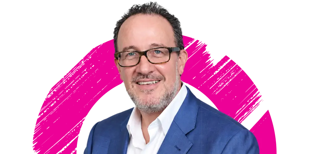 Smiling man with glasses and a beard in a blue suit and white shirt, in front of a white background with a bright pink brushstroke circle.