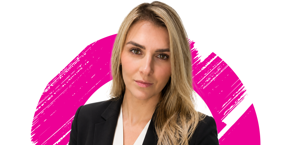 A woman with long blonde hair wearing a black blazer and white blouse stands in front of a white background with a bold, pink brushstroke design.