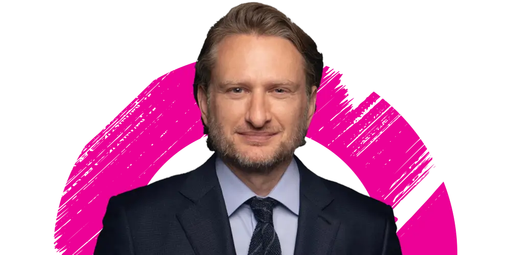 A man in a dark suit and tie smiles slightly, standing in front of a white background with a bold pink brushstroke circle behind him.