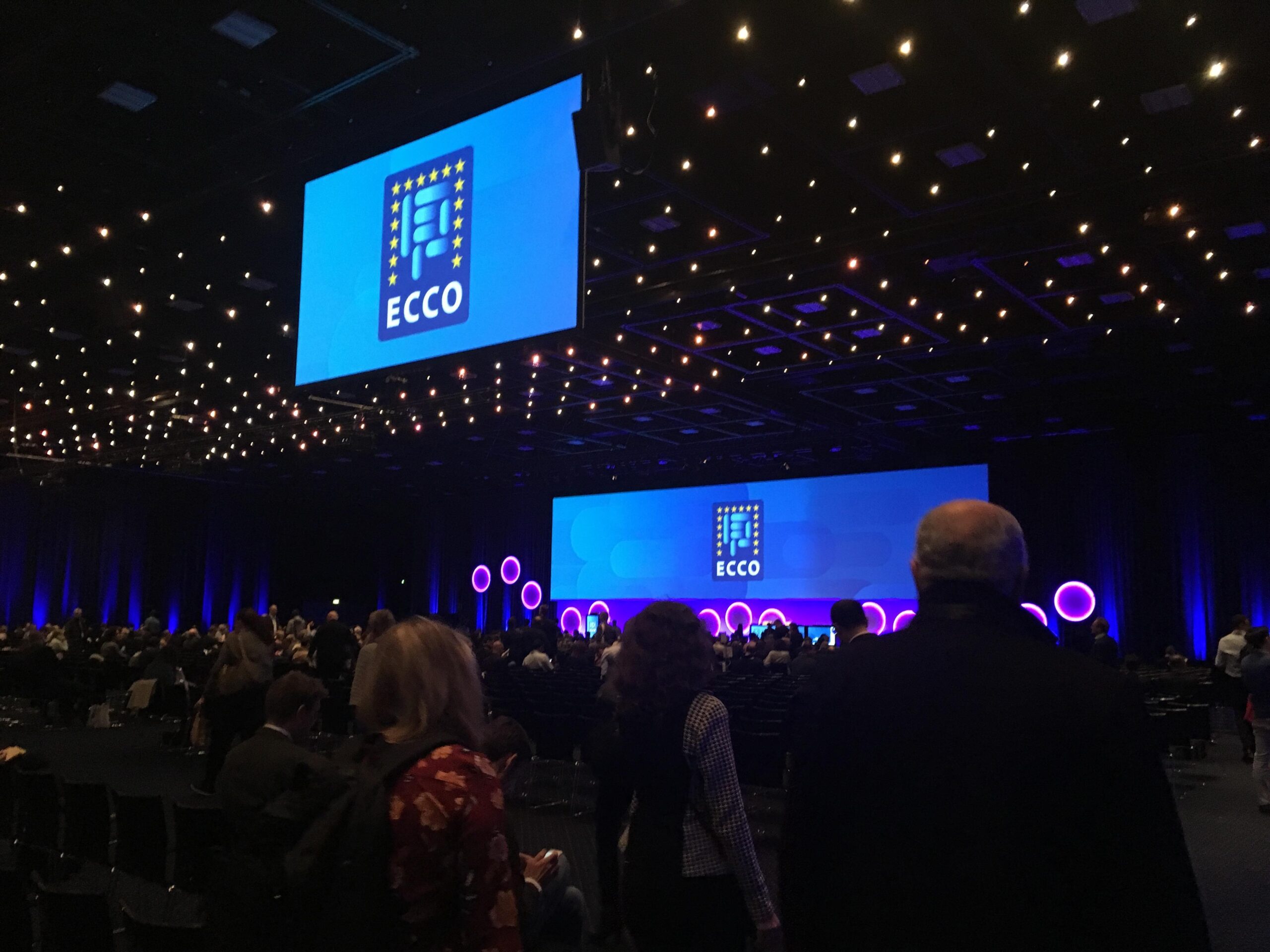 People gather in a large, dimly lit conference hall with blue lighting, circular stage decor, and ECCO logos displayed on two screens. Audience members are seated and standing.